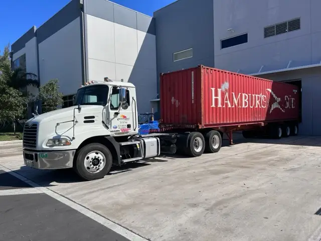 A man steps onto a white semi-truck parked outside a building with a loaded container Drayage companies in miami