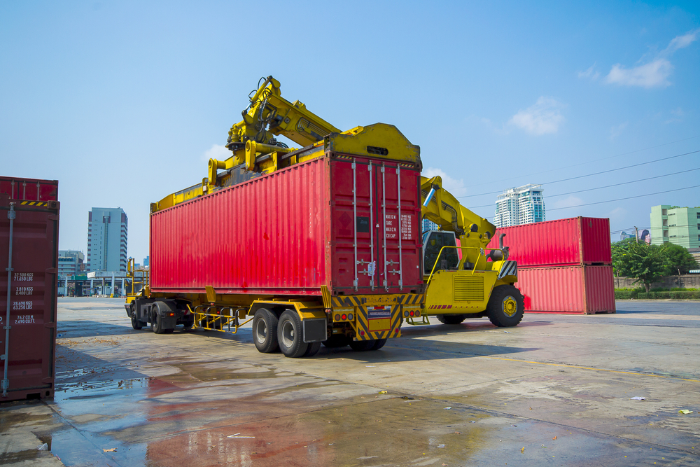 A port worker is using a reach stacker to load a full container onto a chassis trailer for the truck driver to deliver the load